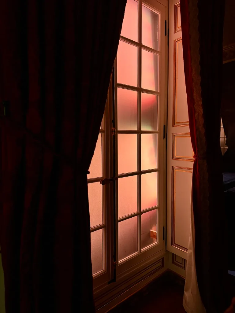 Warm sunset light glimpsed through parted red curtains and a window door. The day self night self essay, 49Grey