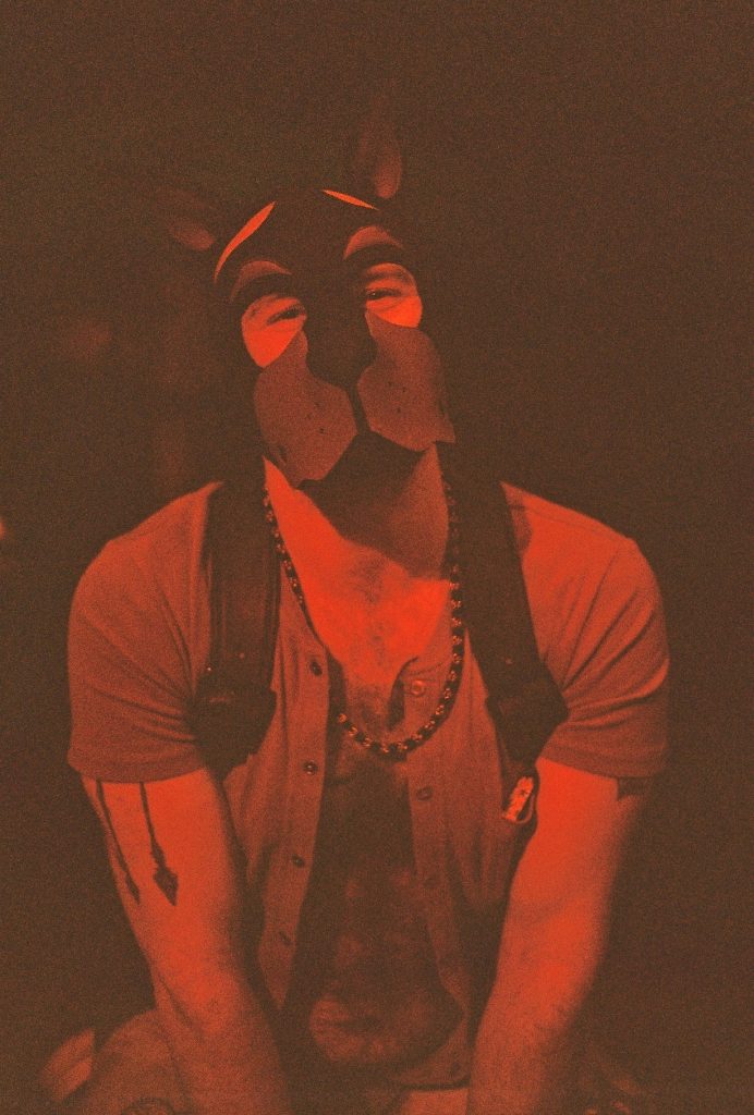Man in pup hood and harness bathed in red light at a leather bar. Pup play essay, 49Grey