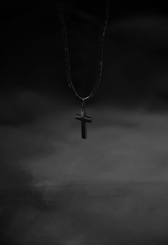 Black cross pendant necklace hanging in dark, moody light — Buffy queer coming out essay, 49Grey