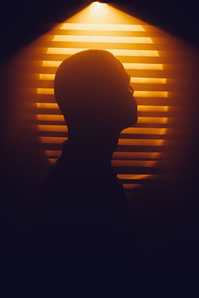 Silhouette of a bald figure lit by warm venetian blind light — George Michael queer icon essay, 49Grey