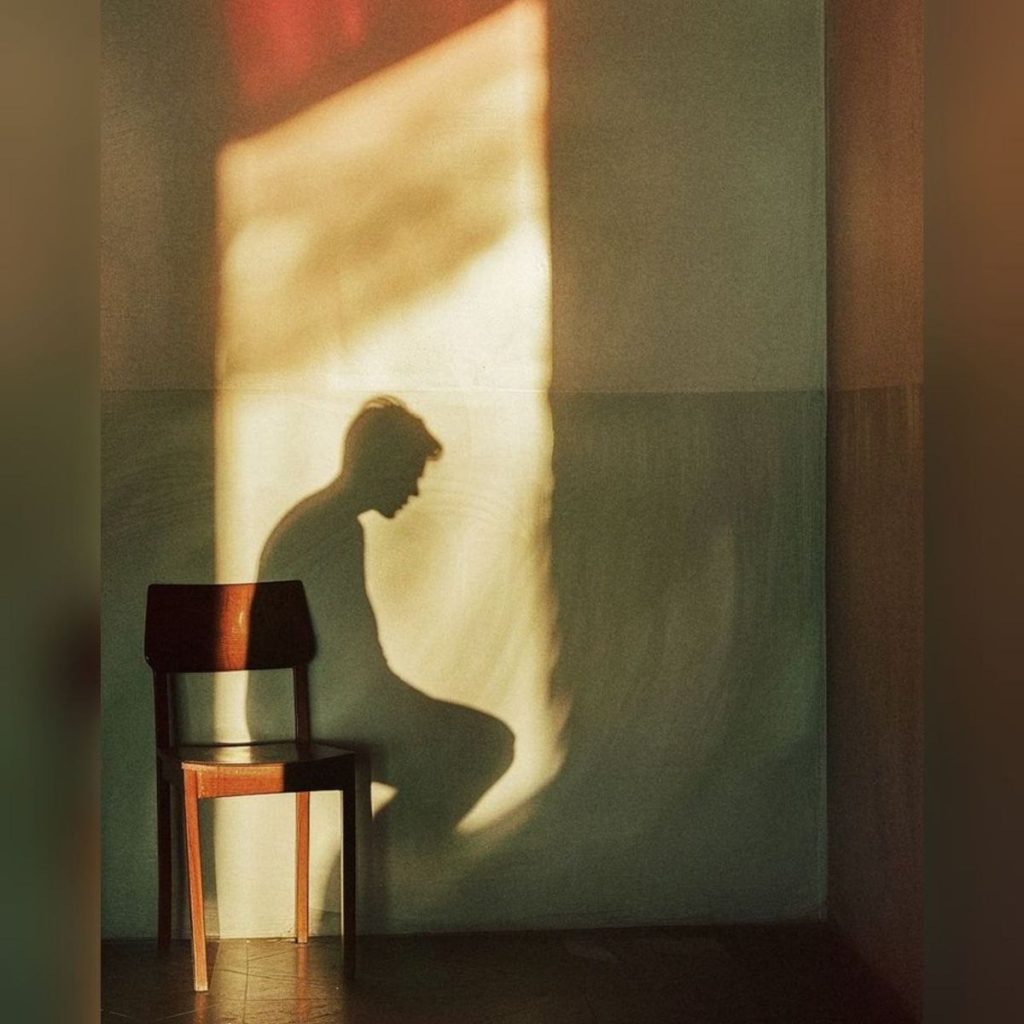 Shadow of a seated figure cast on a wall beside an empty chair in warm light. Shame and desire essay, 49Grey